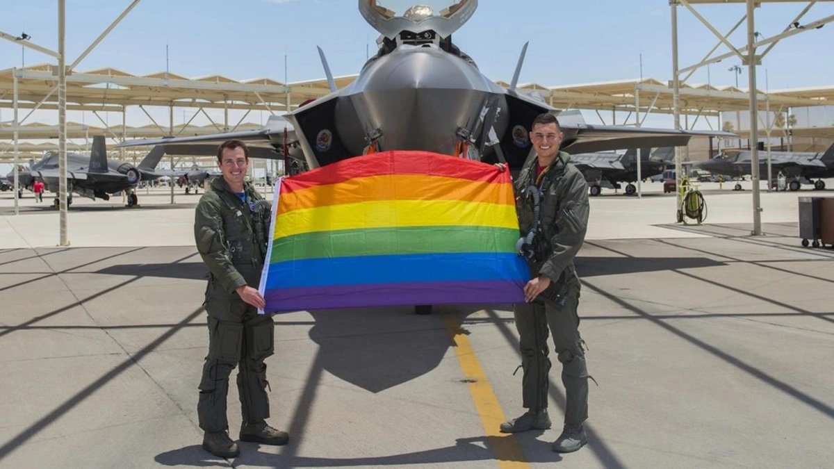 Air Force pilots holding a rainbow flag in front of an F-35 fighter jet on a military base.