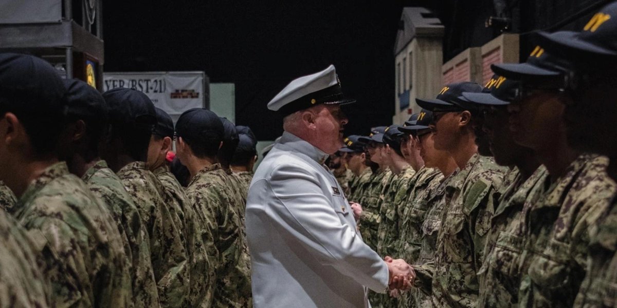 Navy officer in white uniform shaking hands with sailors in camouflage at ceremony.