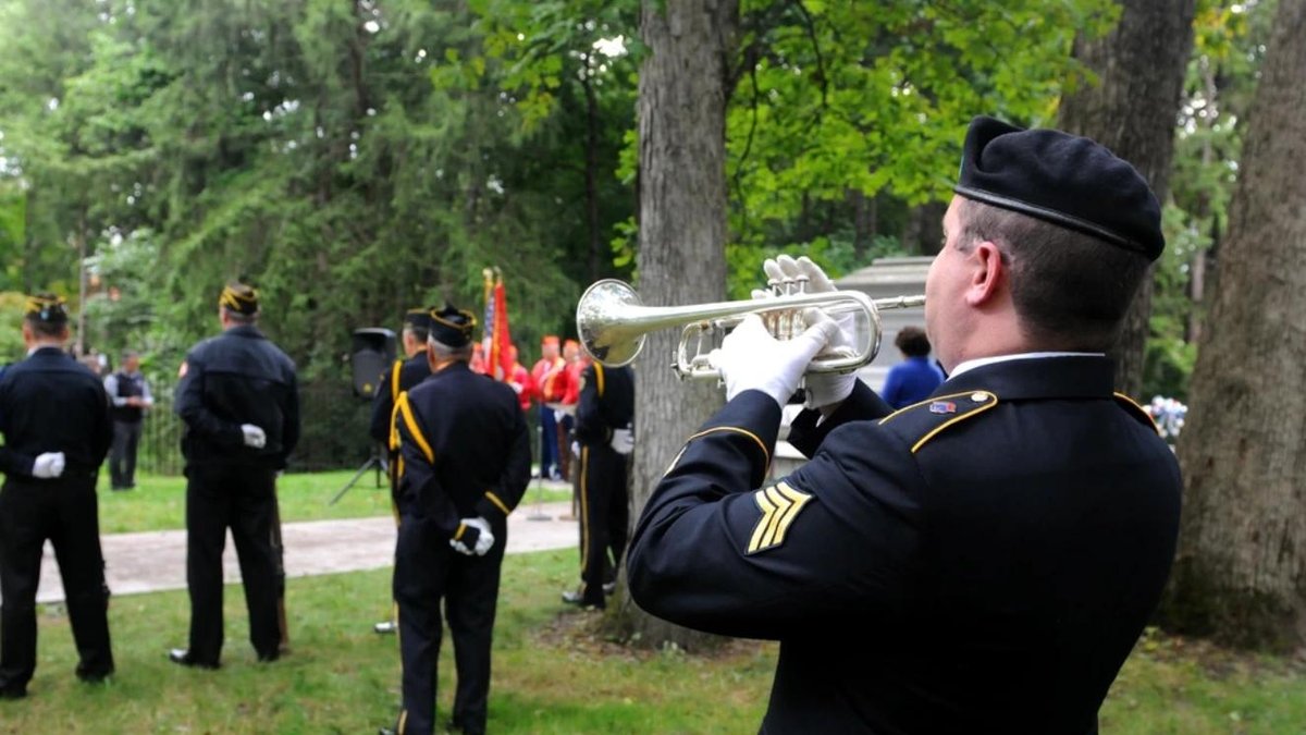 Army bugler playing taps at military funeral ceremony in wooded cemetery setting