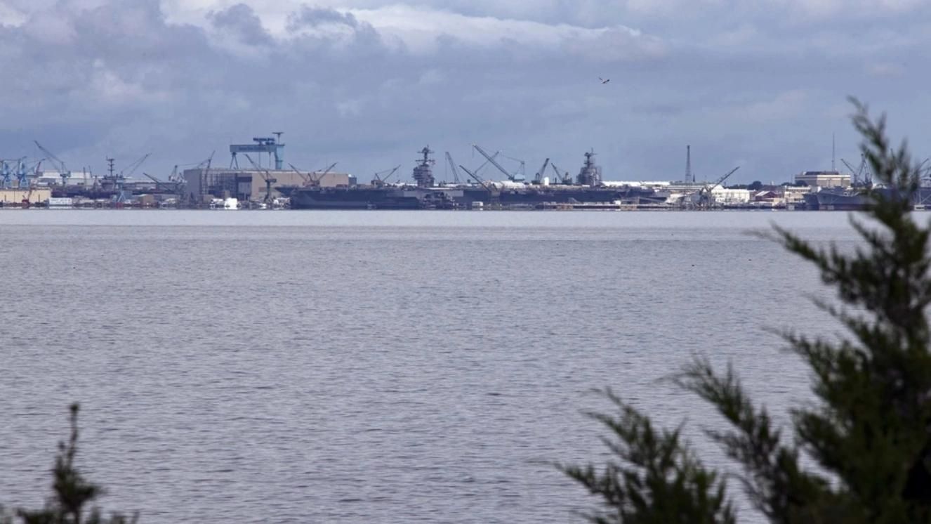 Newport News shipyard with Navy vessels and cranes along the coastline under cloudy sky