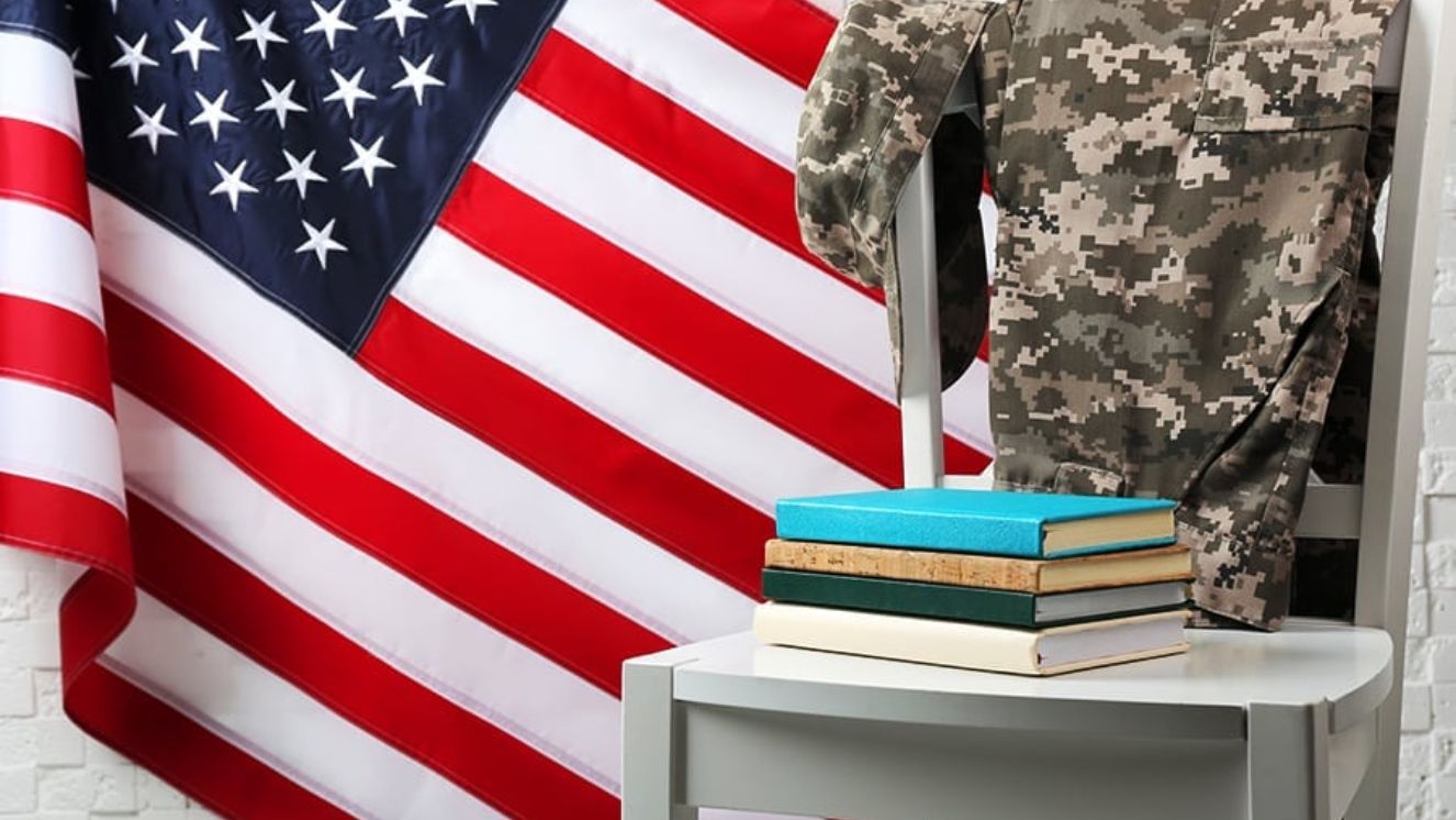 Stack of books on a small table beside a U.S. Army camouflage uniform draped over a chair, with an American flag in the background.