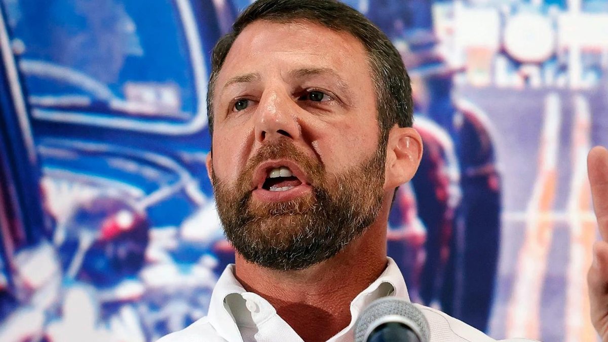 Senator speaking at podium with background of military personnel and vehicles.