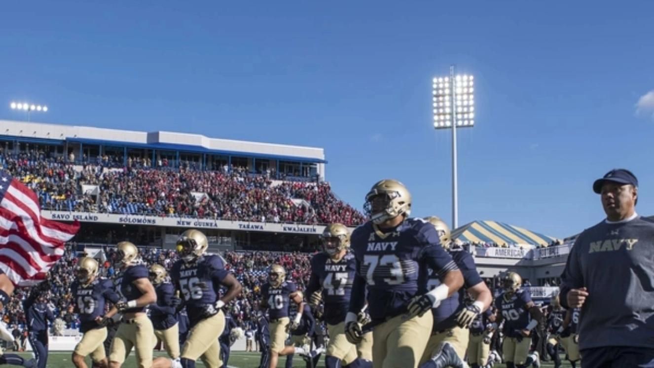 Navy football team entering stadium field under bright lights during game.