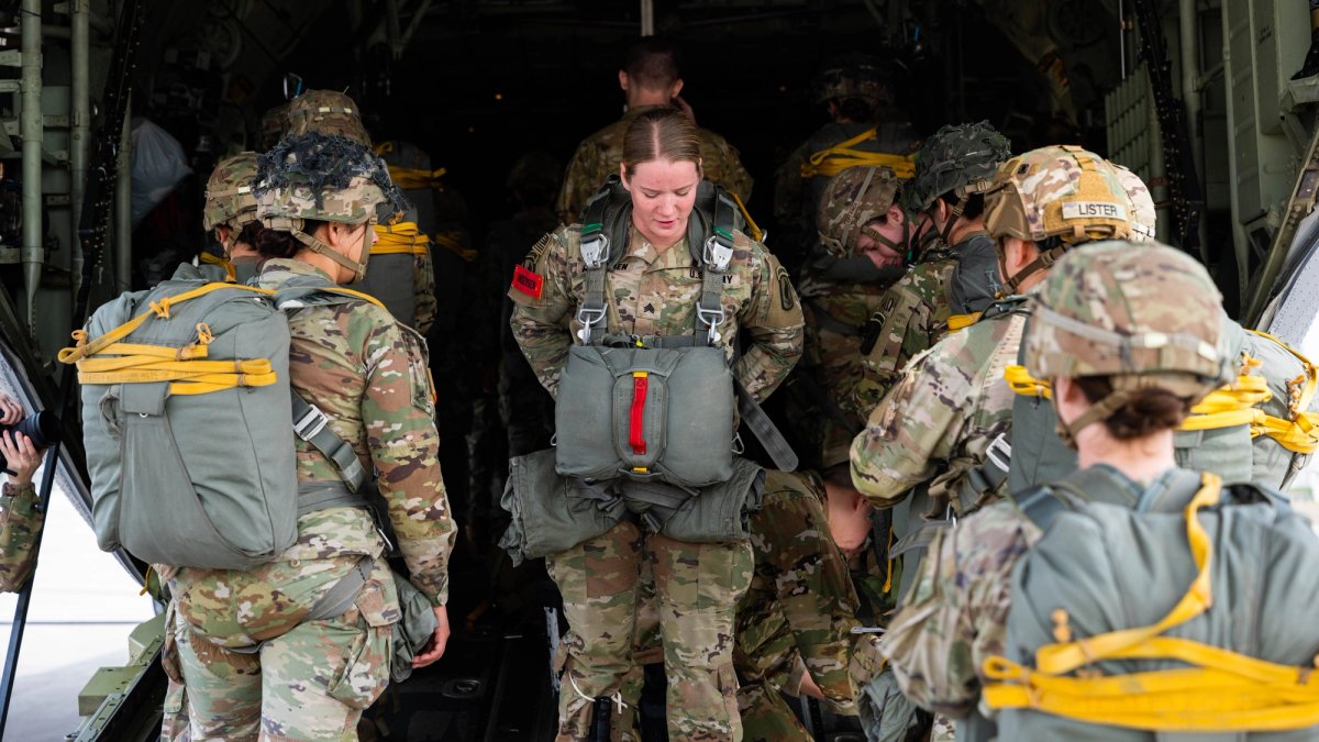 Female paratrooper stands between two rows of soldiers on a plane ramp, adjusting her gear.