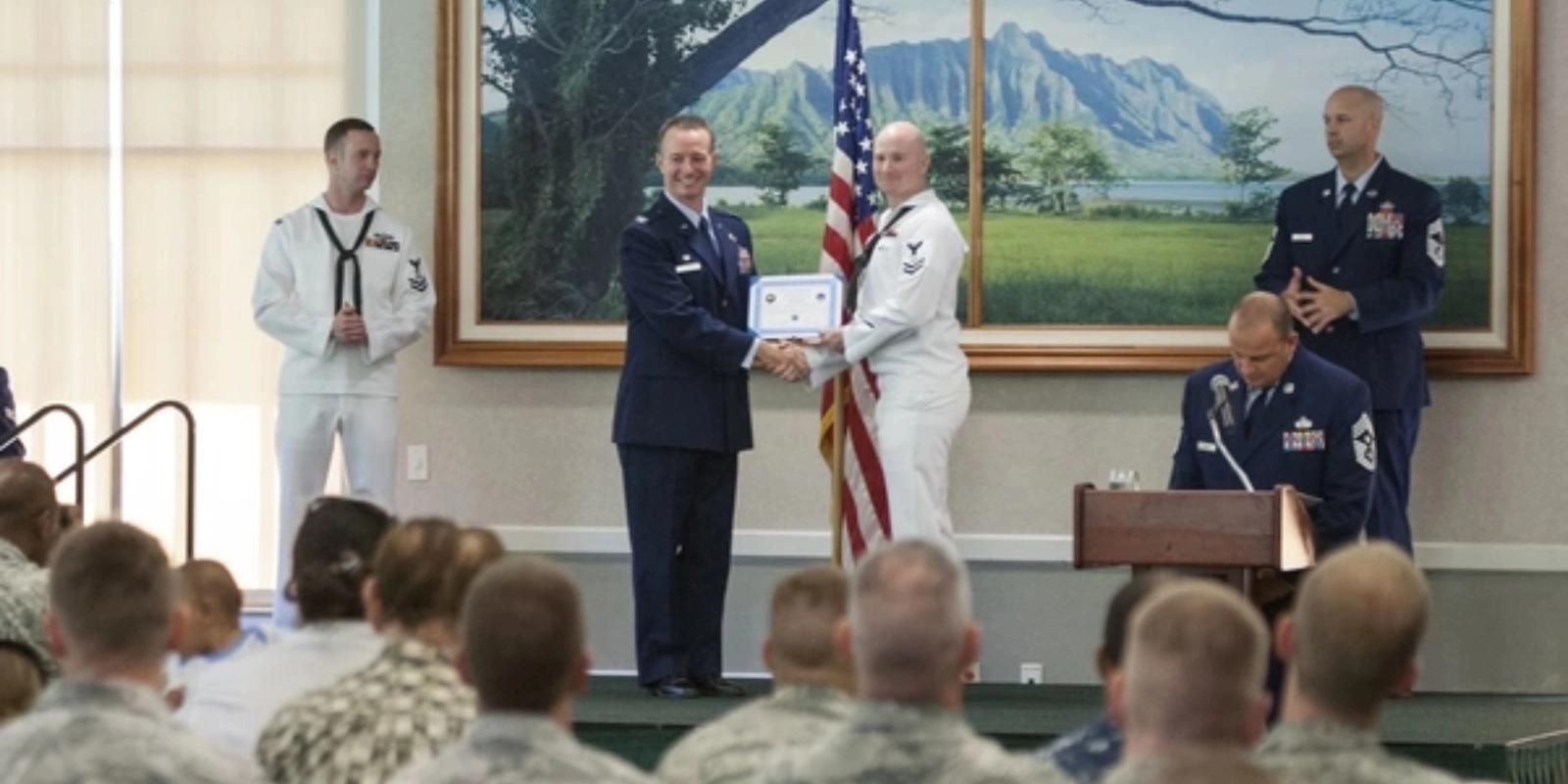 Air Force officer awarding certificate to Navy sailor in ceremony with audience and American flag.