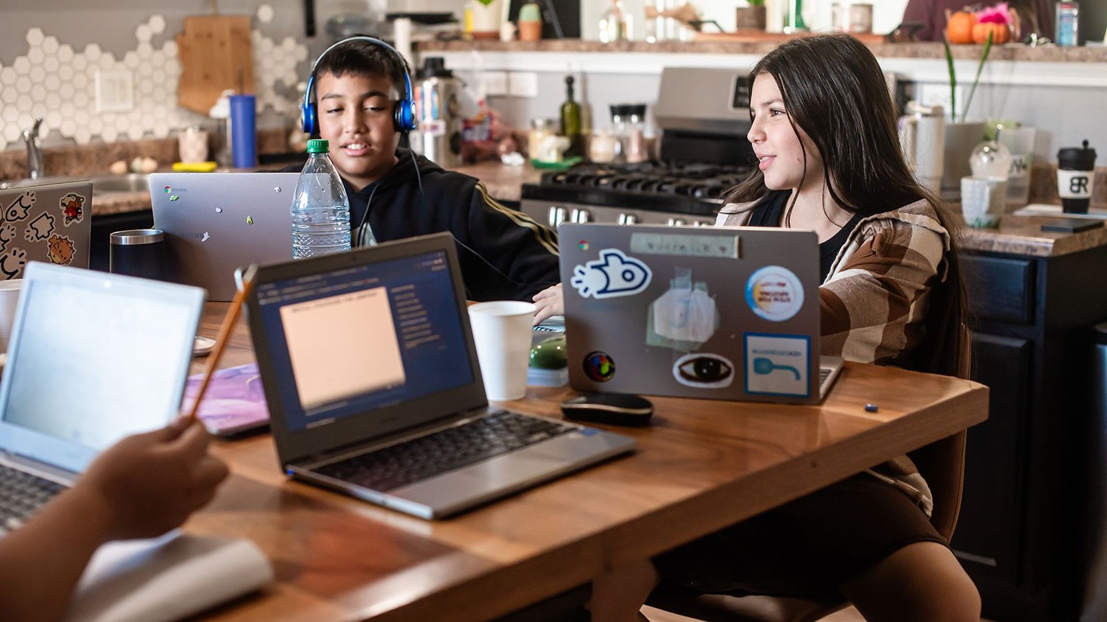 Two students sit at a table with laptops. A hand is seen in the frame holding a pencil.