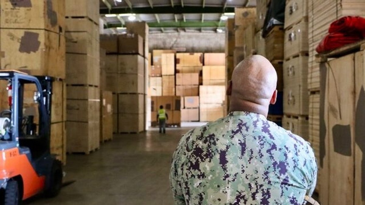 A soldier is seen from behind in a warehouse of moving boxes.