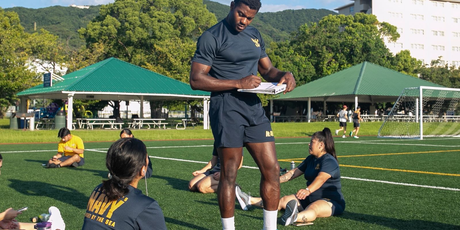 A soldier in a gym uniform records scores of other soldiers performing exercises outdoors on a sports field.