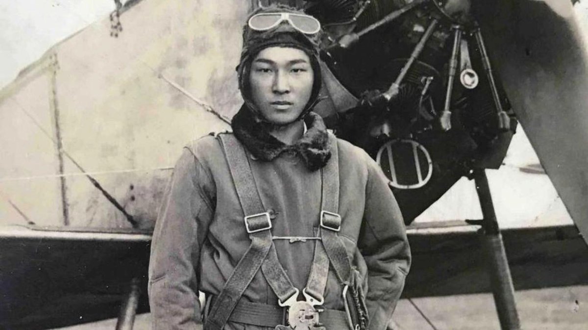 World War II aviator in flight suit standing in front of a vintage military aircraft.
