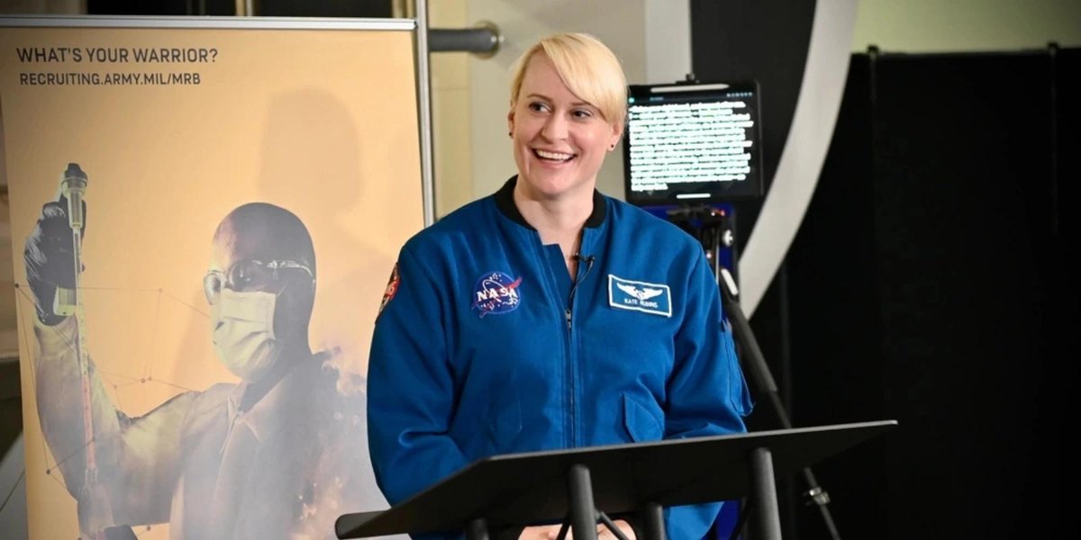 Maj. Kate Rubins in NASA flight suit speaking at podium beside Army recruitment poster.