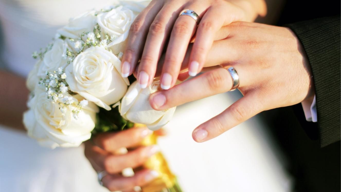 Hands of a married couple with rings holding a bouquet of white roses.