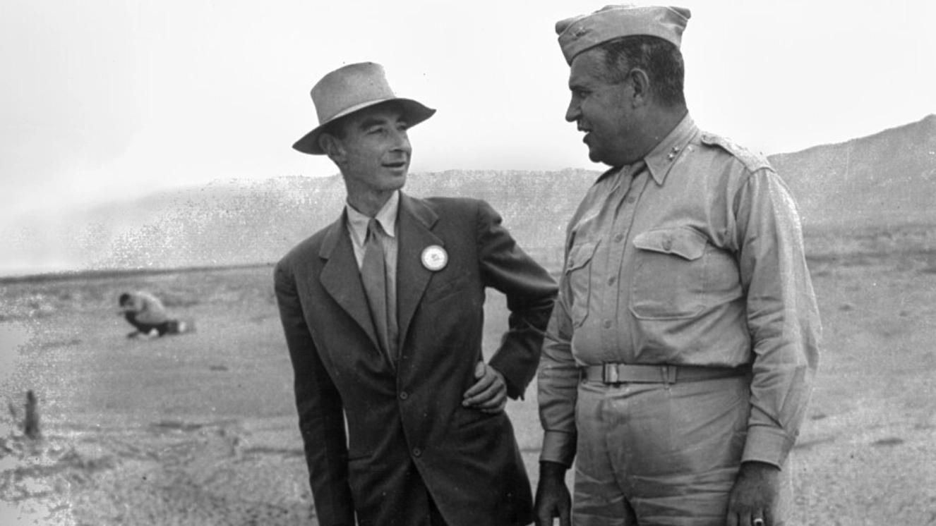 Two men in 1940s attire discussing at a military test site, one in a suit and hat, the other in military uniform.