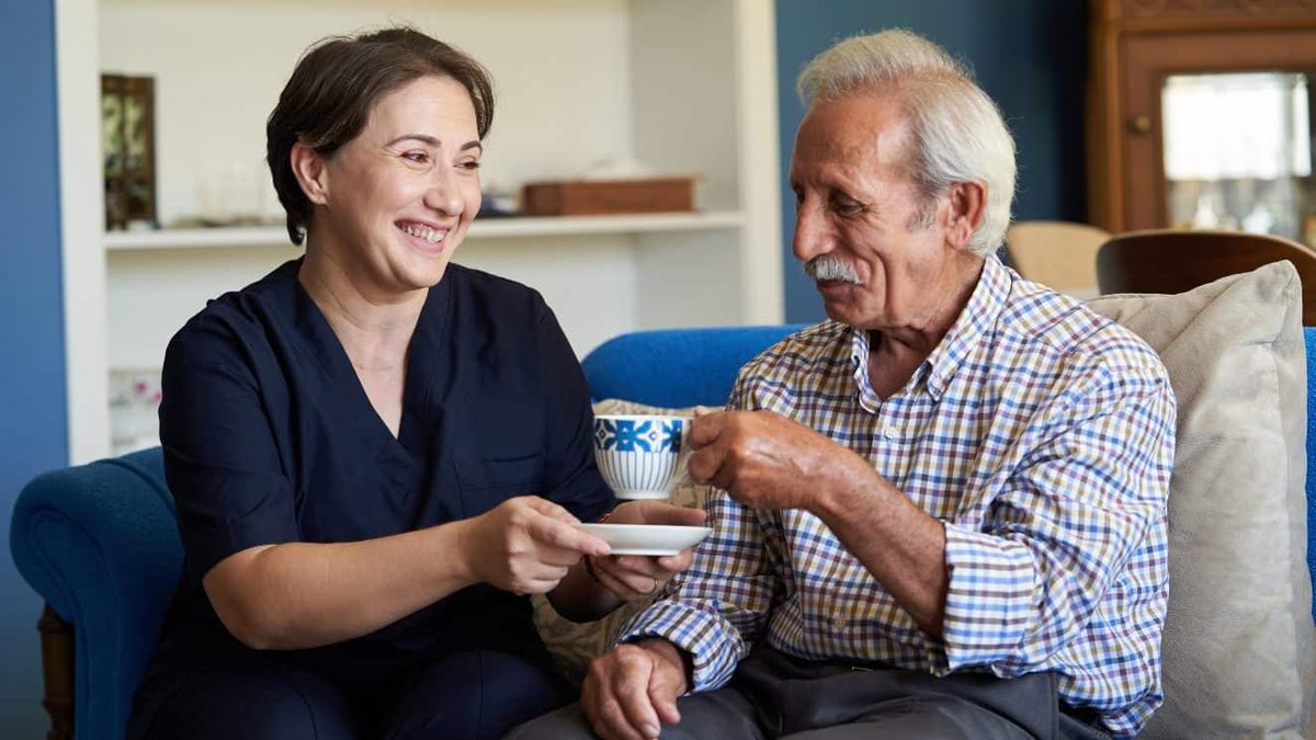 Caregiver assisting elderly veteran with seated tea service in home setting