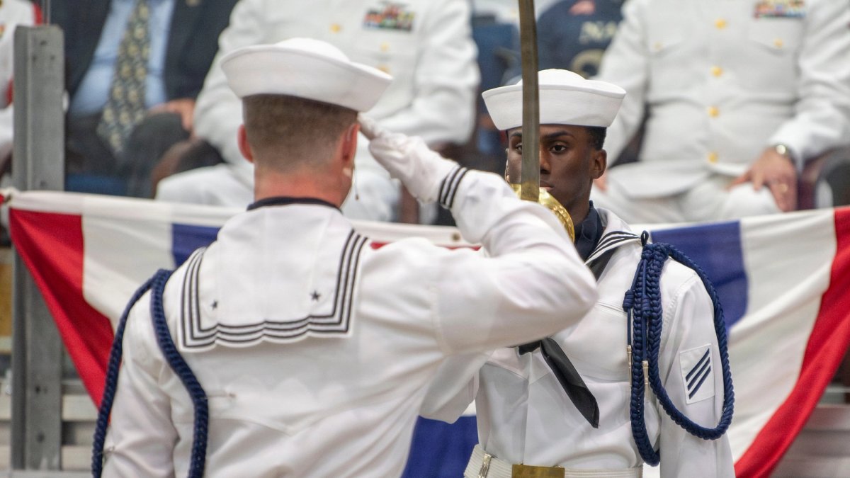 A new Navy recruit salutes another who is holding a sword in front of his face.