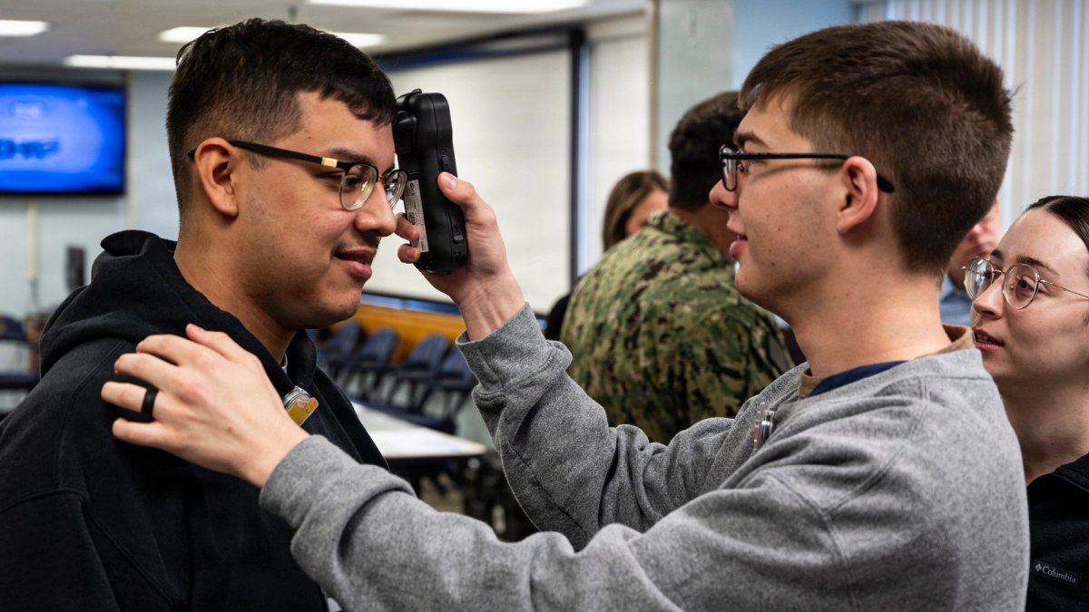 A serviceman holds a device to the head of another serviceman.