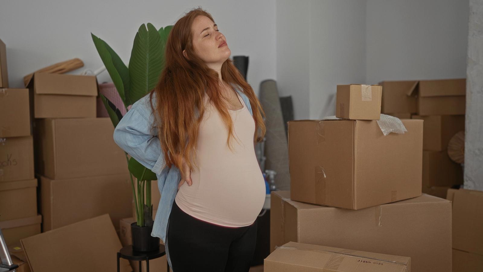 A tired pregnant woman stretches amid moving boxes.