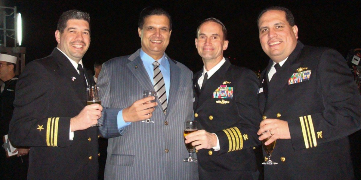 Navy officers in dress uniforms socializing with a civilian man in a suit, holding glasses.