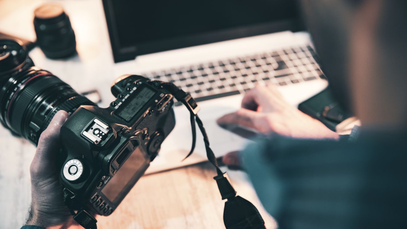 A photographer with a camera in his hand, making edits on a computer at his desk.