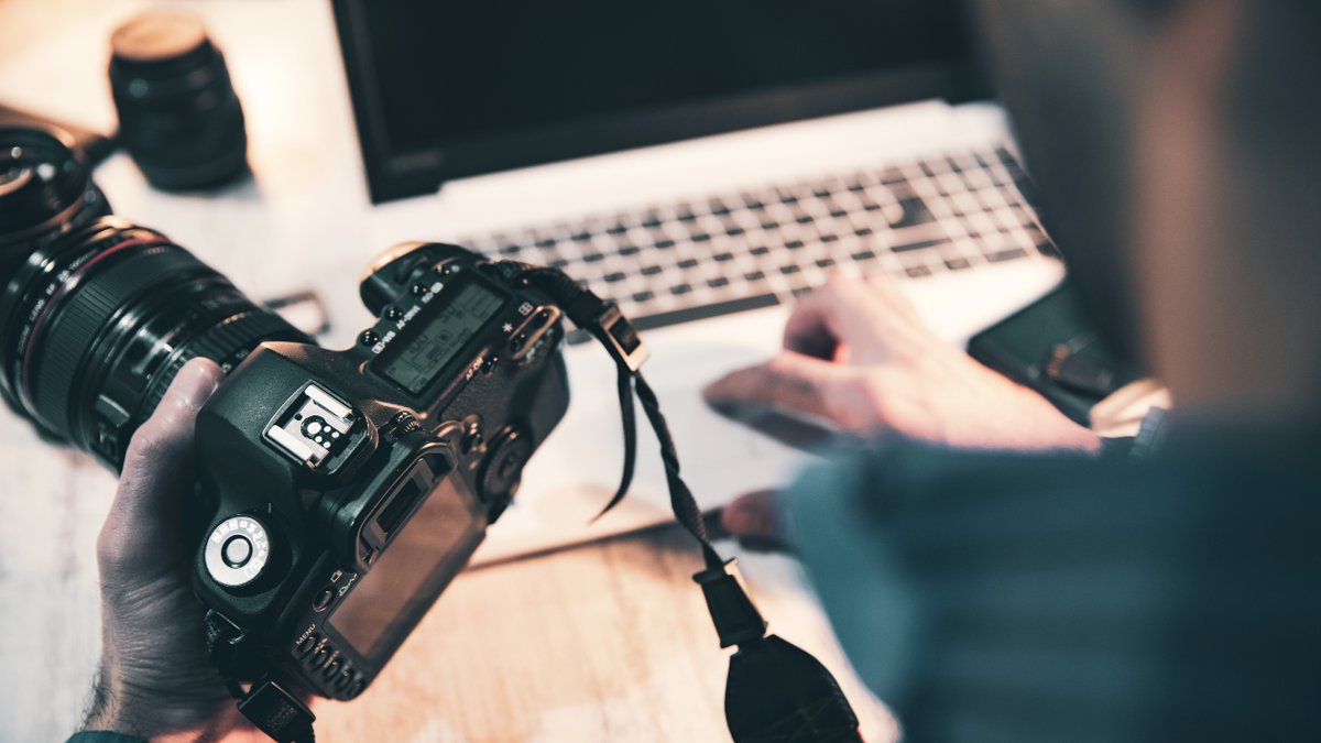 A photographer with a camera in his hand, making edits on a computer at his desk.