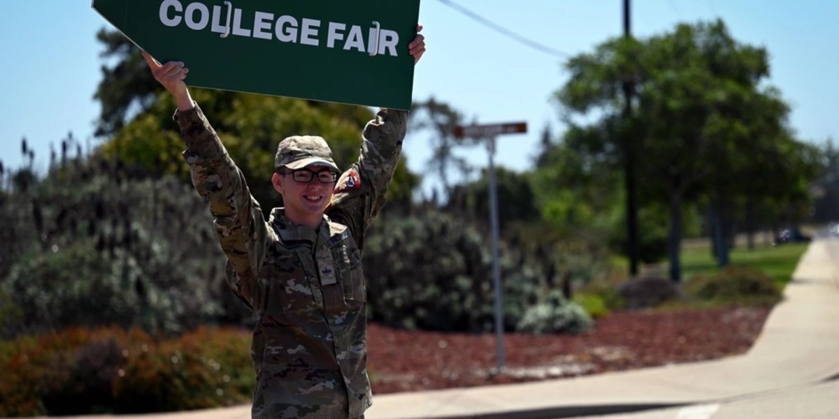 Army soldier in uniform holding a "College Fair" sign outdoors on a sunny day.