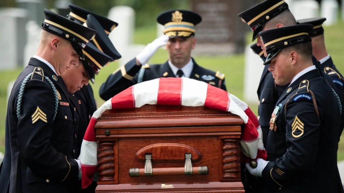 Army soldiers in dress uniforms carrying flag-draped casket at veteran burial ceremony.