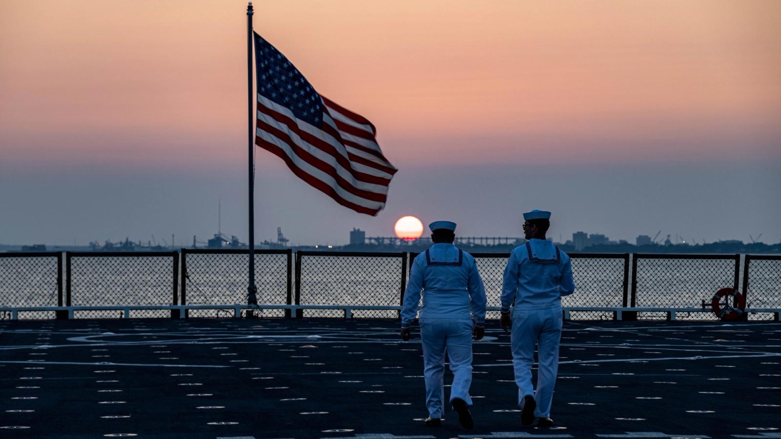 Two sailors on a boardwalk at sunset near a flag.