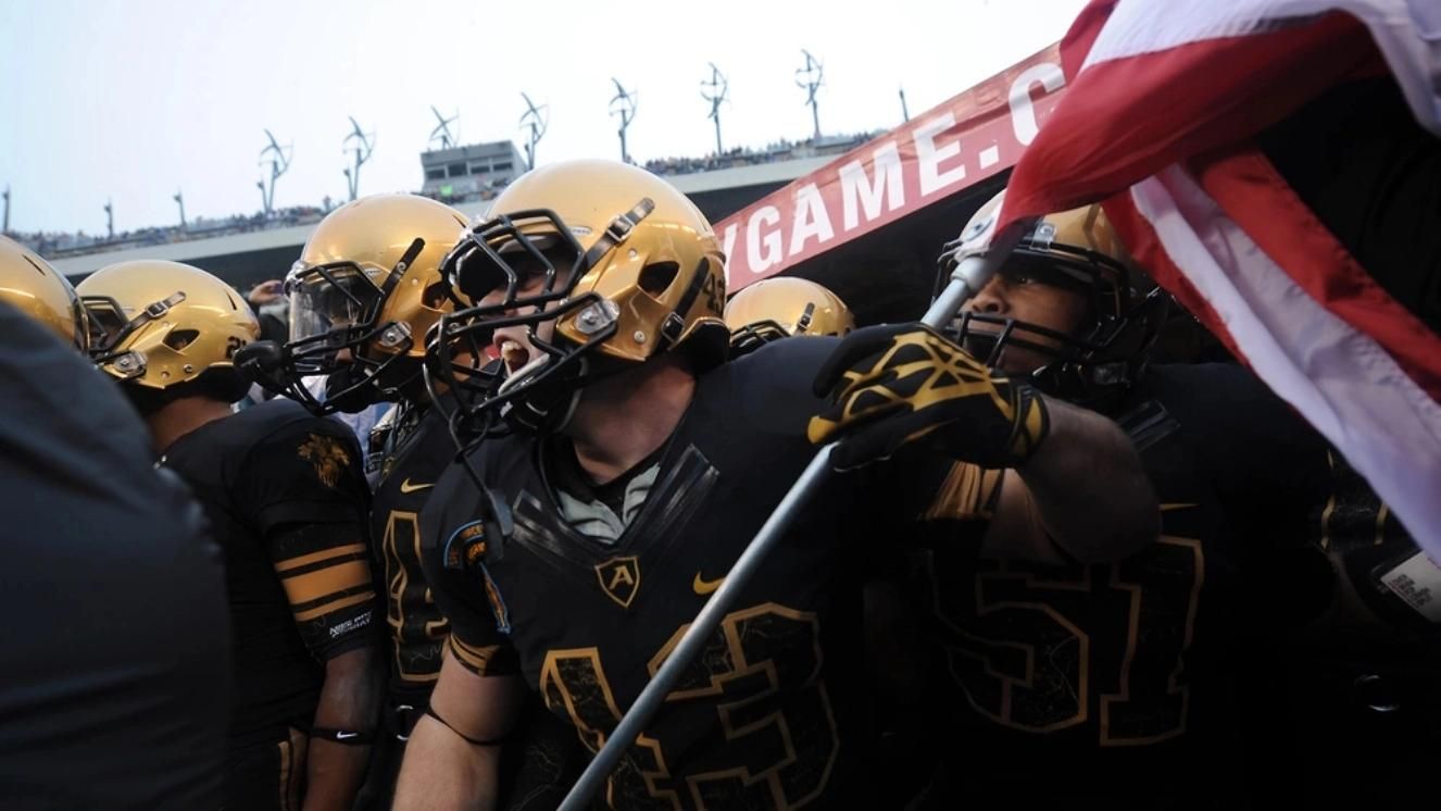 Army Black Knights football players in black uniforms carrying American flag onto field.