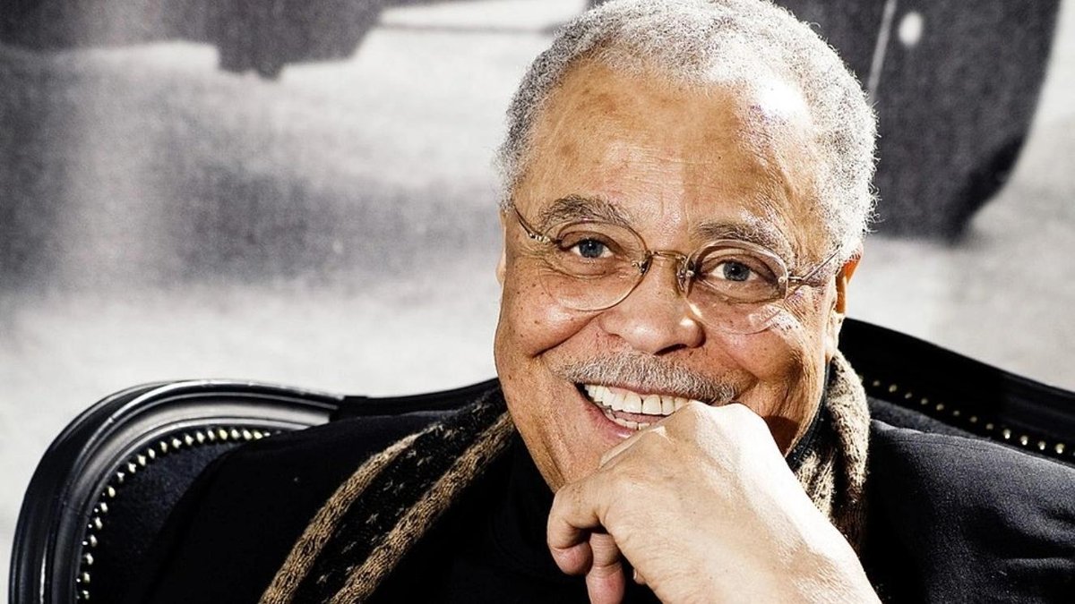 James Earl Jones smiling, wearing glasses and a scarf, seated indoors.