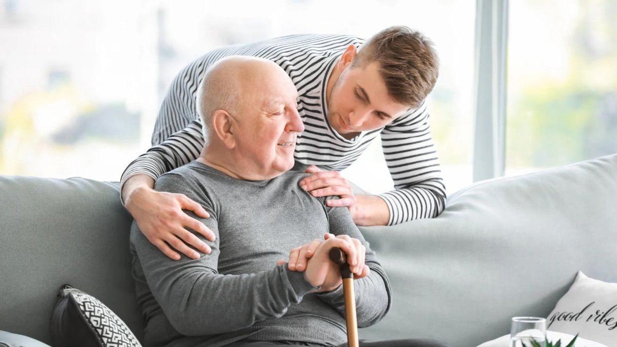 Young man assisting elderly veteran with cane on living room sofa