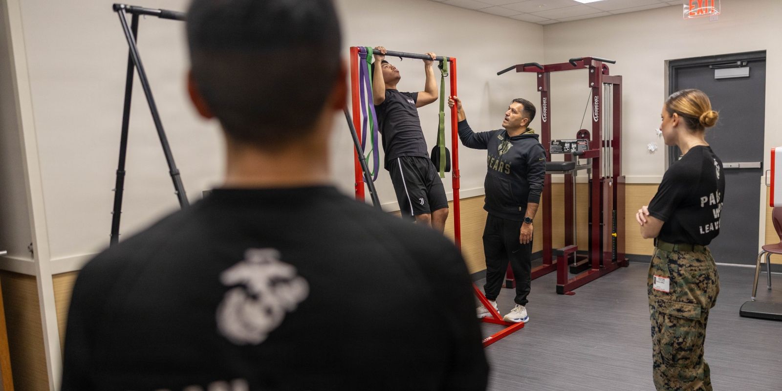 Four servicepeople are in a gym. One man does chin-ups as others watch.