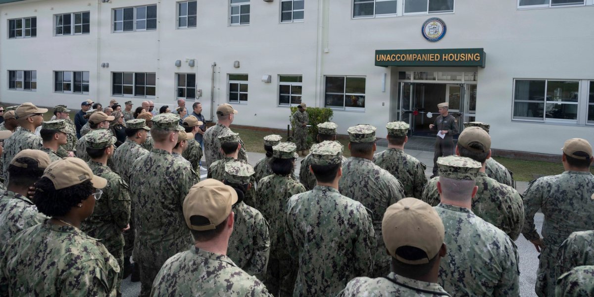 A uniformed serviceman talks to a large group of servicepeople at a ribbon cutting of a new Navy housing unit.