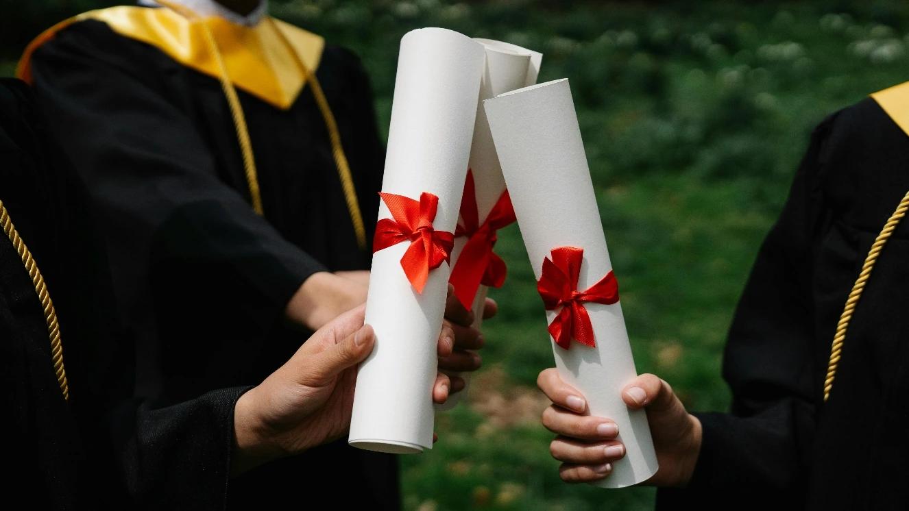 Veterans in graduation gowns holding diplomas with red ribbons outdoors
