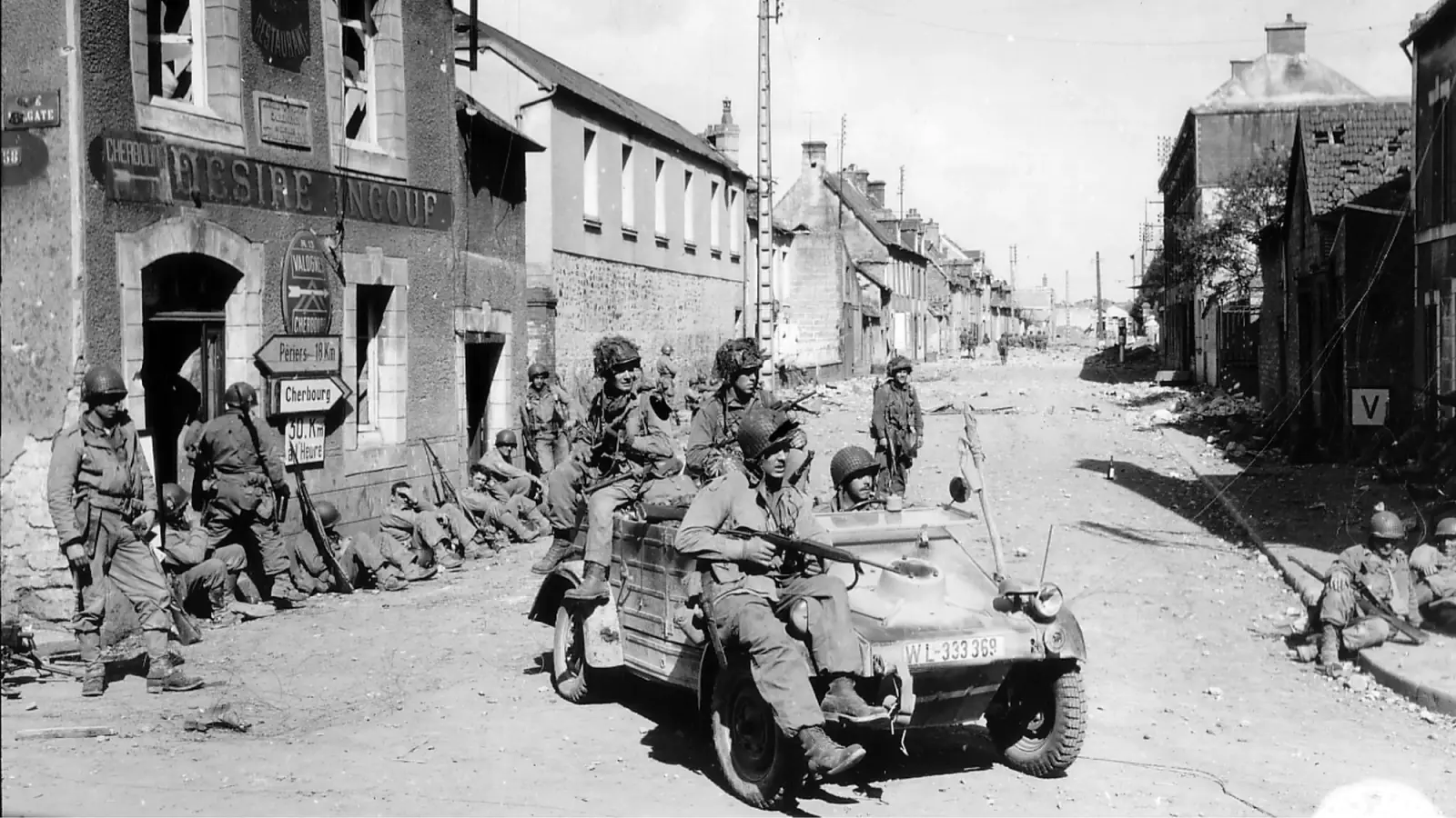 A vintage black and white photo of US soldiers driving through a bombed-out French town.
