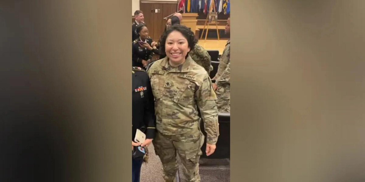 Army personnel in uniform standing indoors with flags in the background.