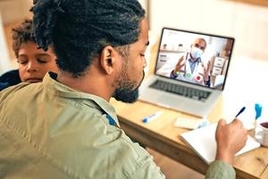 A father talks to a doctor on his laptop while holding a sick child.