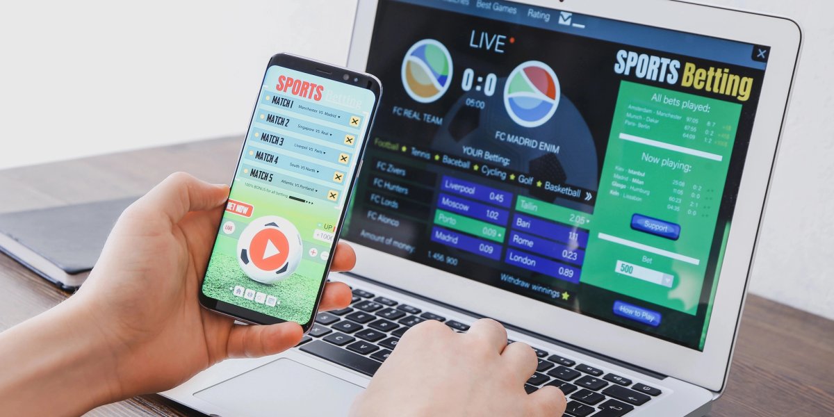 A hand holds a cell phone in front of a laptop with betting websites in view.