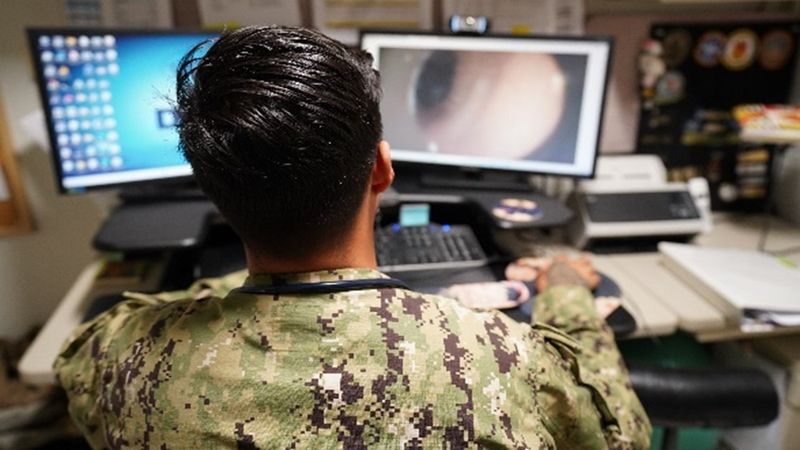 ROTA, Spain. Hospitalman Milo Devore, a corpsman at Medical Home Port, demonstrates a physical assessment using the Telehealth Medical Cart, which enables real-time image sharing for remote provider evaluations. 