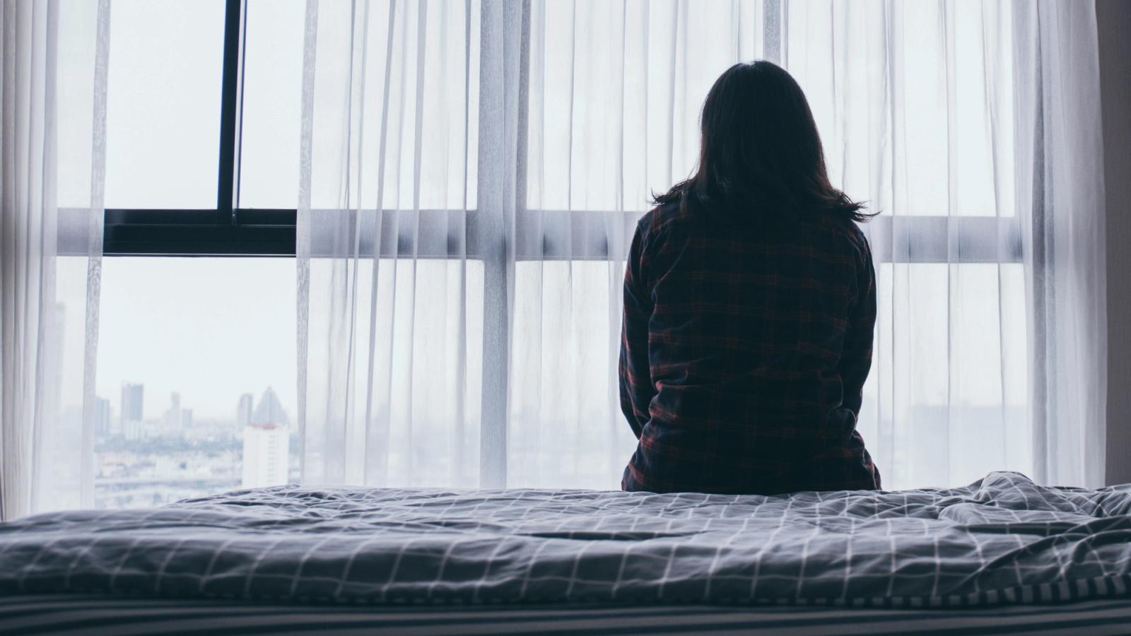 A woman seen from behind staring out of a window with sheer curtains.