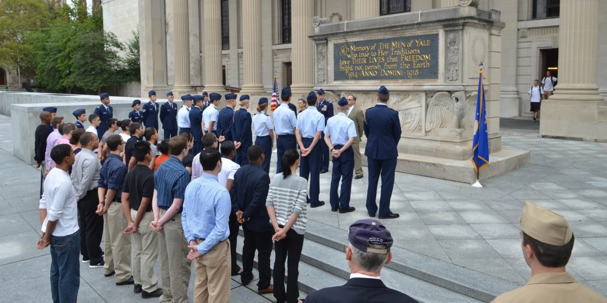 A group of students stands outside in front of a plaque at Yale, listening to a speaker.
