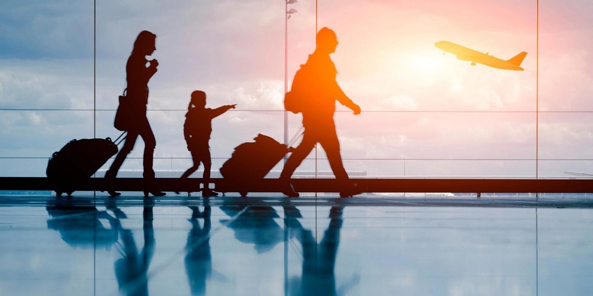 Silhouetted family with luggage walking in airport terminal as plane takes off.
