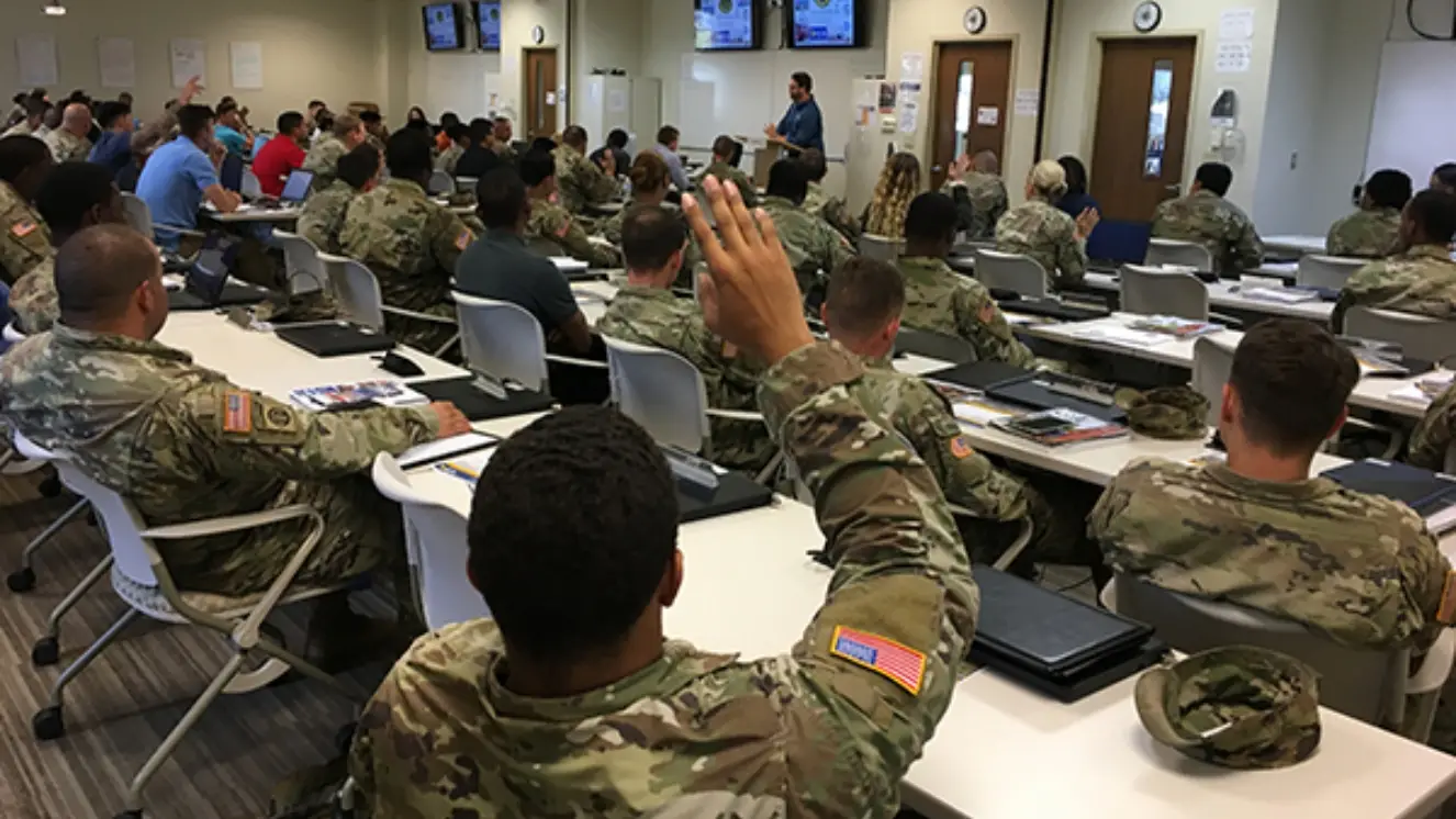 Uniformed soldiers sit in a classroom with a hand raised.