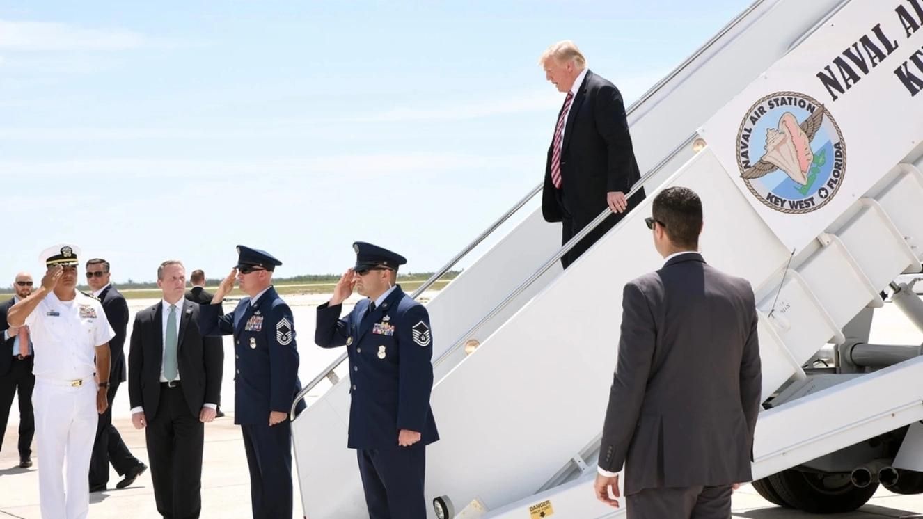 Trump descending stairs of Air Force One at Naval Air Station Key West, greeted by military officers.