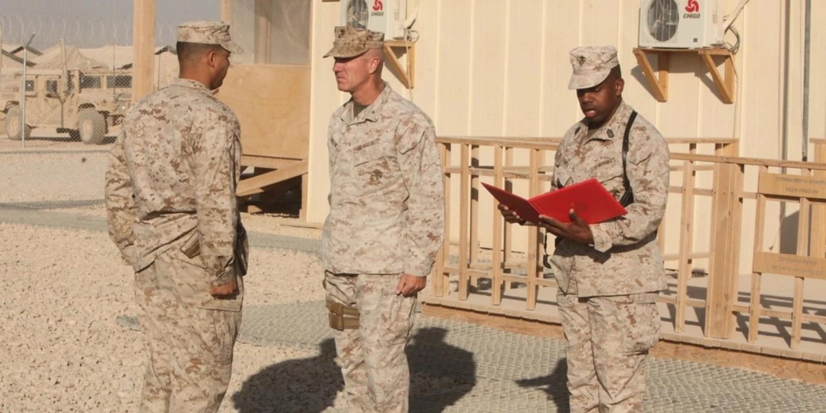 Marine Corps officers discussing outside a desert base building with one holding a red folder.