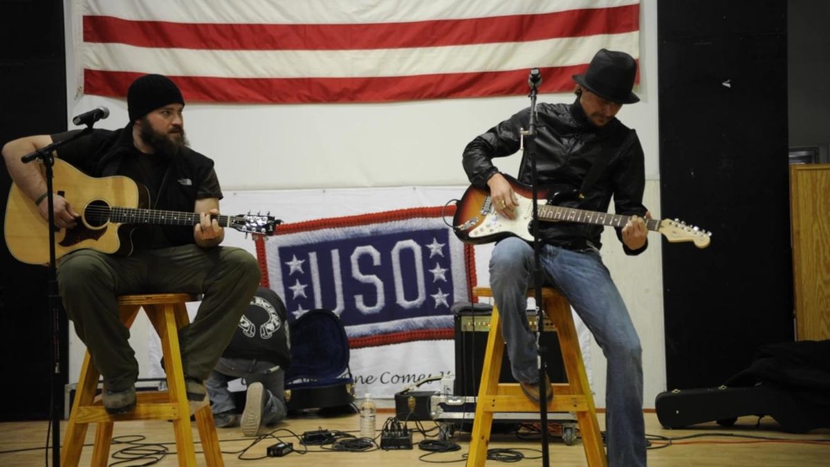 Musicians performing at USO show with guitars, American flag backdrop, USO banner visible.