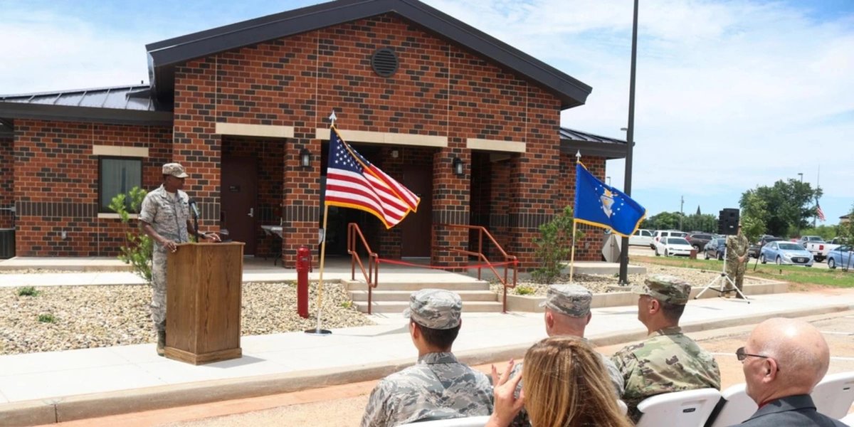 Air Force officer speaking at base ceremony with US and Air Force flags in front of brick building