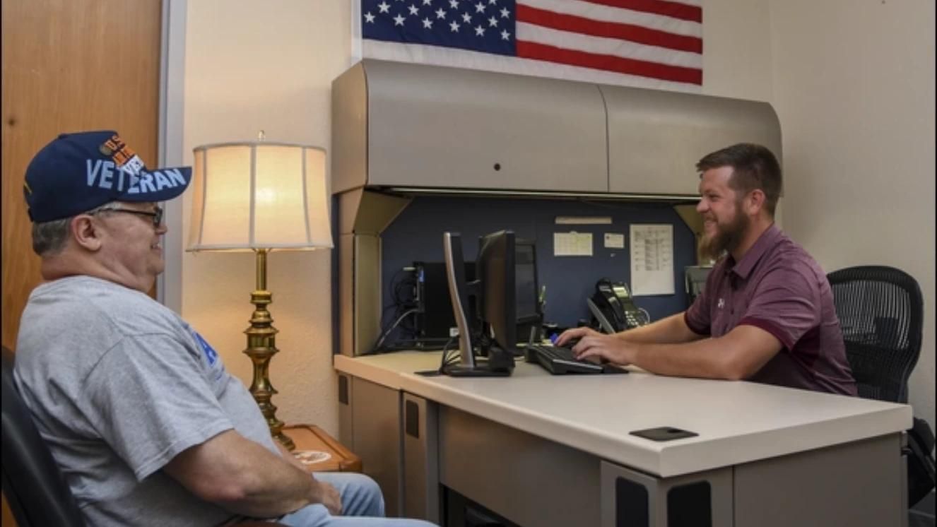 Veteran discussing pension rates with VA representative in office under American flag.