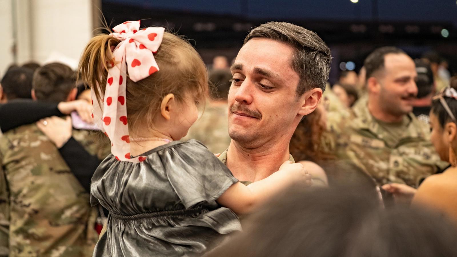 A uniformed Dad looks at his daughter in his arms.
