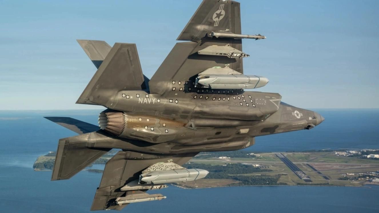 Navy F-35 jet in flight over coastal airbase with AGM-158C LRASM missiles