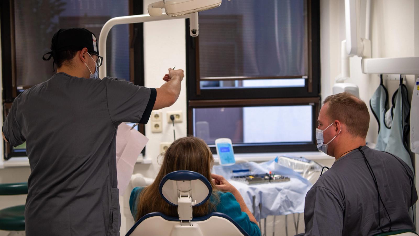 A dentist and dental technician sit on either side of a patient seen from behind.