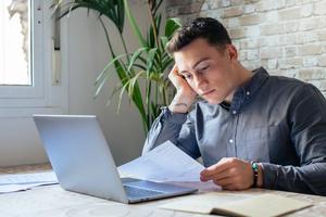 Young man with a laptop looking at a form very seriously.
